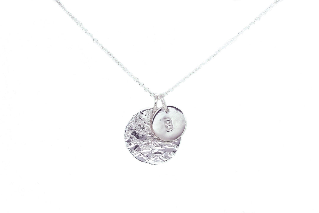 A delicate silver necklace with a chain holds two circular pendants. One pendant has a textured surface, while the other is smooth with the letter 'B' hand stamped on it. The background is white, making this Personalised Large Disc Charm in Silver by Personalised Charms and its sterling silver charms the focal point.