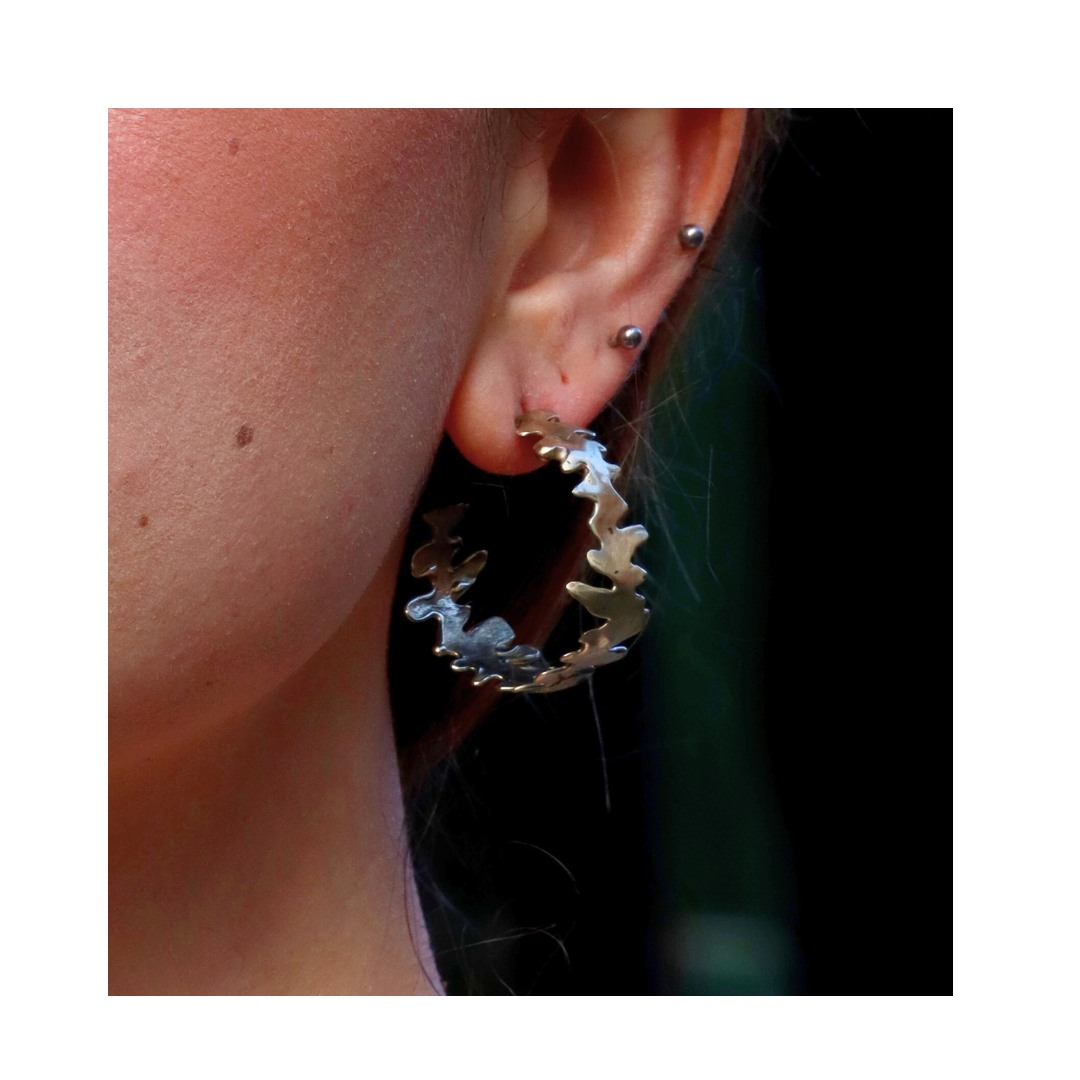 A close-up of a person's ear with multiple piercings showcases the Lava Hoops by Madeleine Sculptural Jewellery, featuring large, jagged silver hoops with organic lines. A dark, blurred background highlights their unique design.