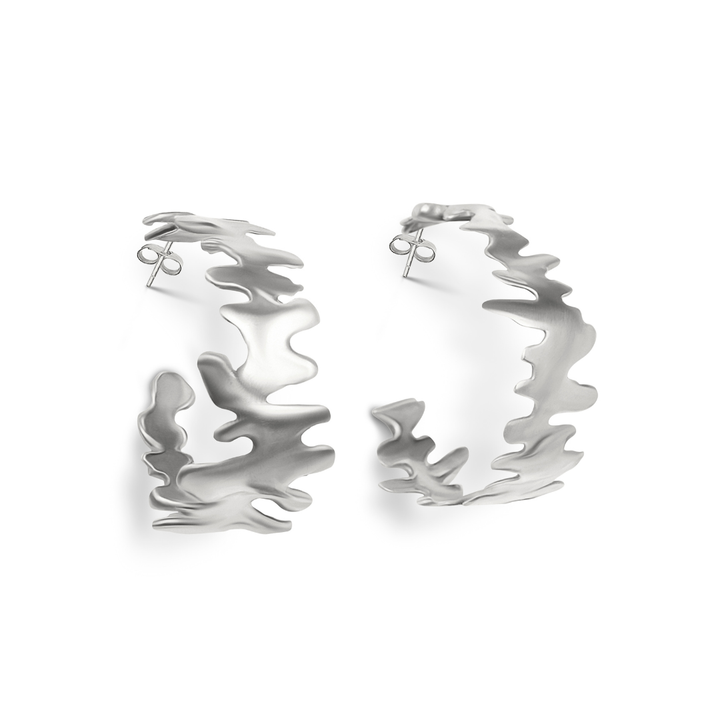 The Lava Hoops by Madeleine Sculptural Jewellery are handmade silver hoop earrings with a wavy, abstract design and matte finish, shown against a white background—an elegant statement piece.
