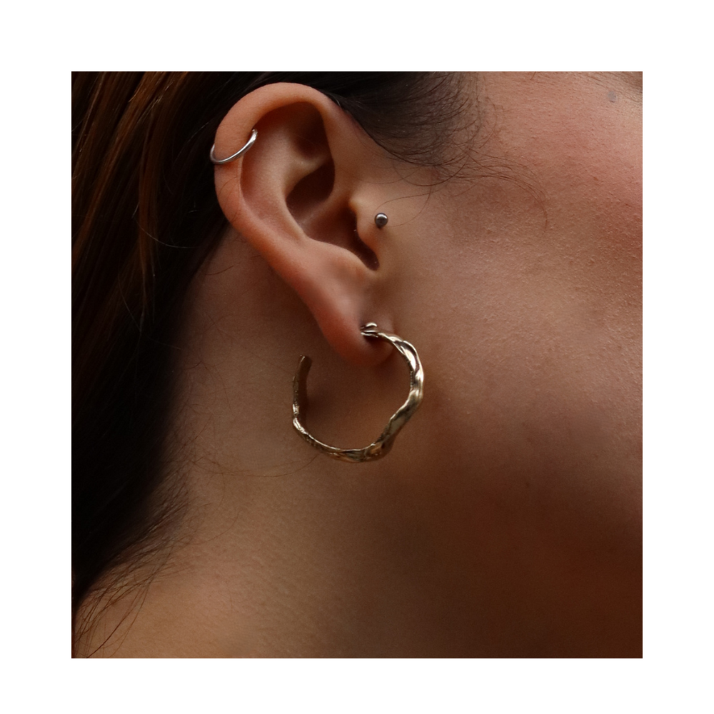 Close-up of a person's ear adorned with three earrings: a small hoop on the upper ear, a stud in the middle, and the Liana Hoop—a large, hand-carved gold earring from Madeleine Sculptural Jewellery—on the lower lobe. The plain white background highlights this beautiful piece of Dublin-designed jewelry.
