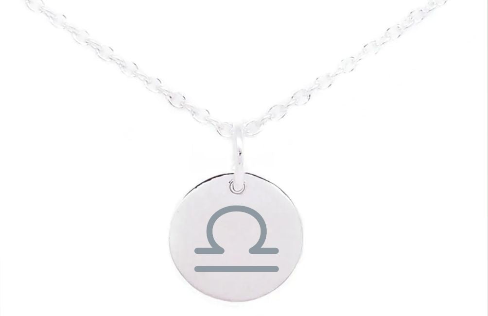 A silver necklace with a delicate chain features a round pendant engraved with the Libra zodiac symbol. The Horoscope Large Disc Charm Silver, part of our Personalised Charms collection, hangs centrally from the fine, intertwined links of the chain. The plain white background accentuates the necklace's elegant design.