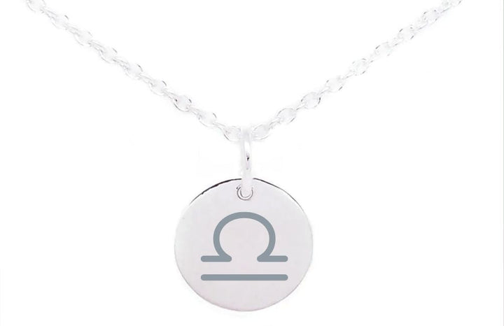 A silver necklace with a delicate chain features a round pendant engraved with the Libra zodiac symbol. The Horoscope Large Disc Charm Silver, part of our Personalised Charms collection, hangs centrally from the fine, intertwined links of the chain. The plain white background accentuates the necklace's elegant design.