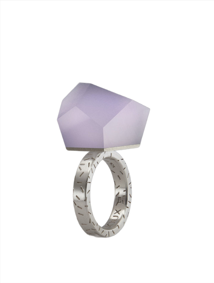 The Fruit Bijoux Fruit Bijoux Ring Top VU Crystals Lilac Purple is topped with a large, asymmetrical, faceted lavender gemstone. The ring has a modern and unique design, with crisp lines and a contemporary aesthetic.