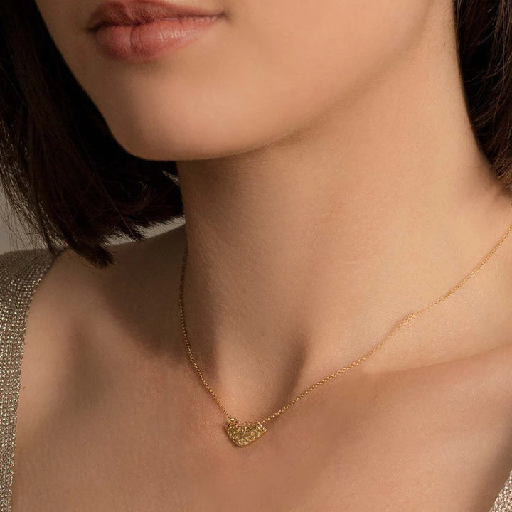 Image showing a close-up of a woman’s neck and collarbone area, focusing on a delicate gold plated sterling silver necklace called the Little Shield Necklace Gold from Andrea Mears Jewellery with an intricately designed heart-shaped pendant. She is wearing a subtle shimmering top with a V-shaped neckline. Only a portion of her lips and chin are visible.