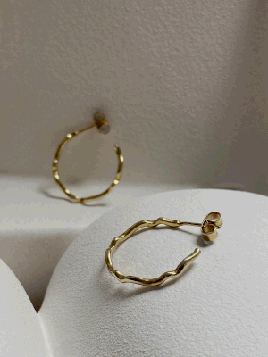 The Sister Hoops by Inner Island, featuring a wavy, textured gold design, are elegantly displayed on a white curved surface—perfect versatile earrings for jewellery lovers. One hoop lies flat while the other stands upright in the background.