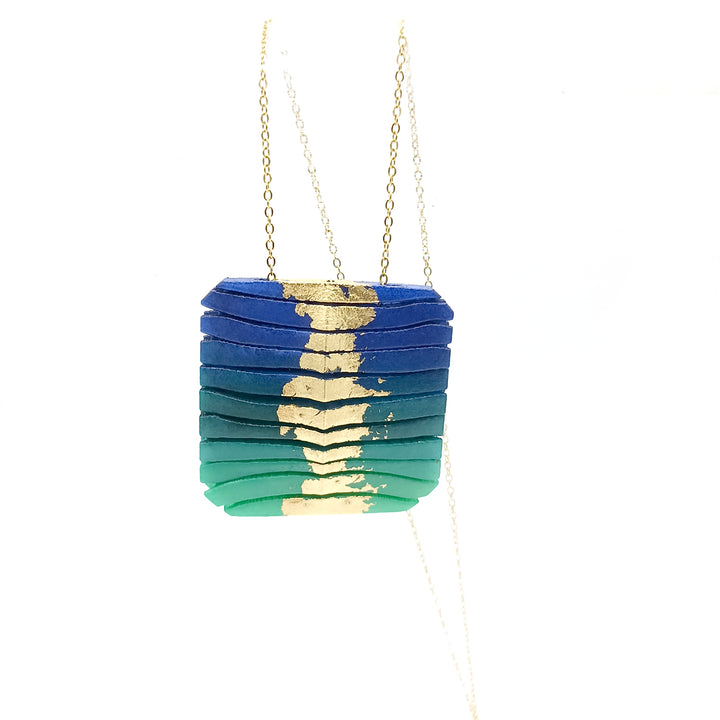 A Tochu-De Long Square Wave Necklace in Blue & Emerald, featuring a pendant with a horizontal layered design, transitioning from dark blue at the top to green at the bottom. Each layer is adorned with gold accents in the center. The pendant, crafted from powder coated 3D printed nylon, is suspended from a delicate gold chain length 28".