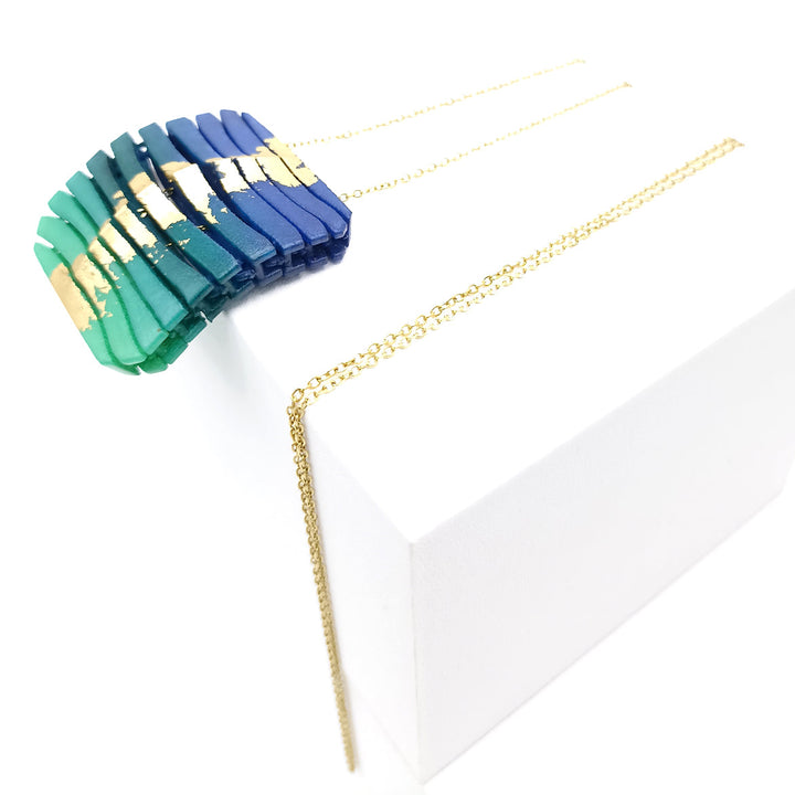 A modern Tochu-De Long Square Wave Necklace in Blue & Emerald featuring rectangular resin pieces in gradient shades of blue, teal, and green with gold accents. The pieces are arranged in a fan shape, attached to a delicate gold chain. With a chain length of 28", the necklace is displayed on a white platform with trailing chains.