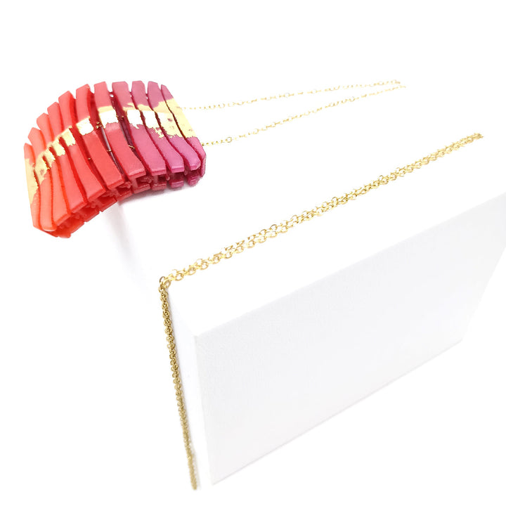 A Tochu-De Long Square Wave Necklace in Fuchsia & Orange with a series of vibrant, rectangular beads in shades of red, pink, and orange, arranged in a gradient pattern, is displayed on a white box. The delicate gold chain adds elegance while careful jewellery care ensures its shine and prevents oxidation over time.