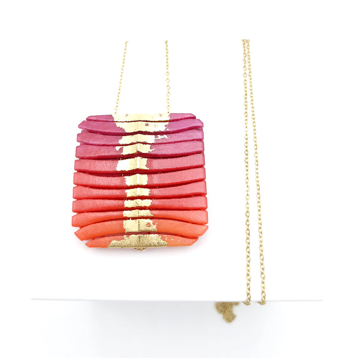 The Long Square Wave Necklace in Fuchsia & Orange by Tochu-De features a pendant composed of layered, thin rectangular pieces arranged in a stack. The 3D printed nylon pieces vary in color from deep pink at the top, graduating through shades of red to bright orange at the bottom, with a gold accent running vertically through the center.