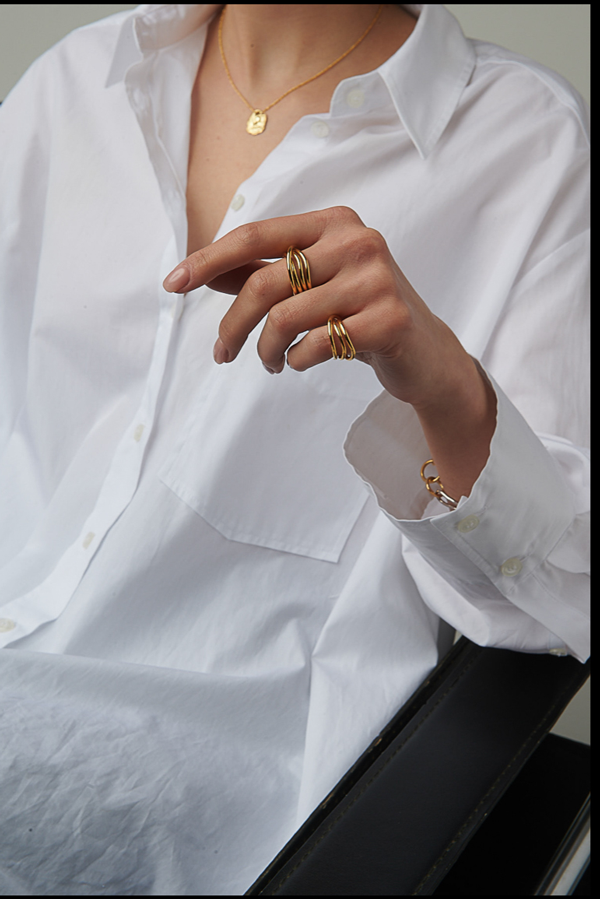 A person wearing a white button-up shirt is shown from the chest down. They are accessorized with a gold necklace and multiple Loop Rings by Bonvo on their fingers. The hand is elegantly positioned, and they are seated in a black chair.