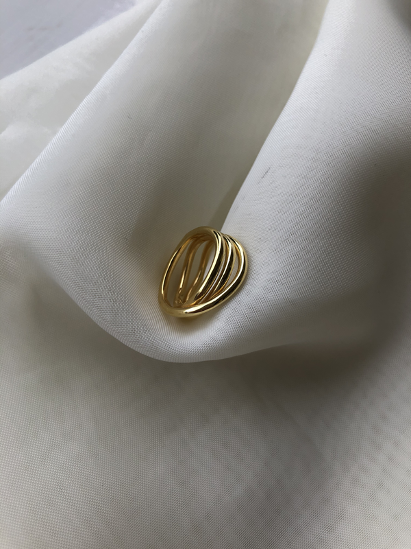 A Bonvo Loop Ring with a sleek, modern design featuring multiple intertwined bands rests on a soft white fabric backdrop. Crafted with 18ct gold plating, the delicate curves of the ring contrast elegantly with the smooth, flowing texture of the fabric.