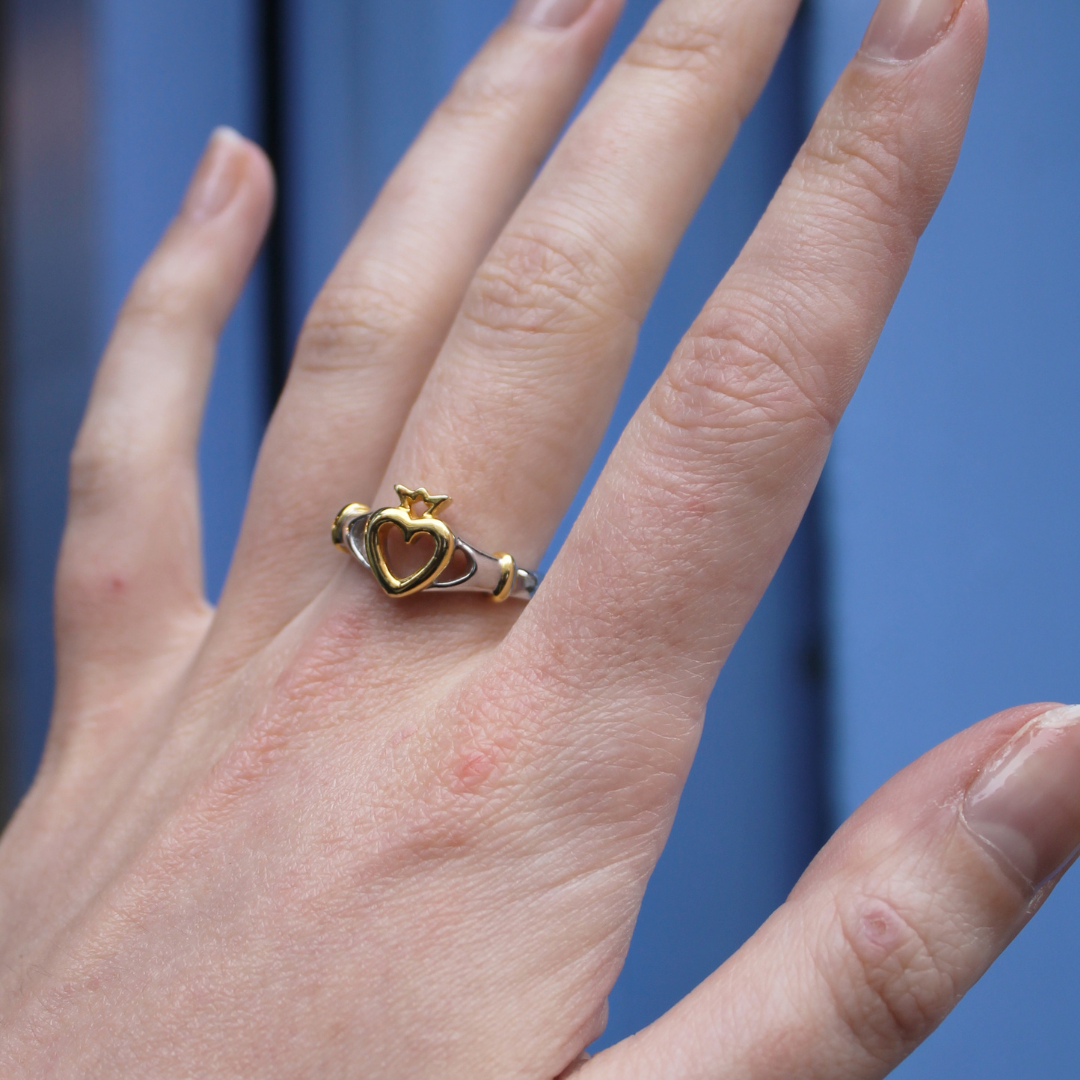 A hand with natural, unpolished nails wears the Watermelon tropical Lorien Ring— a sterling silver Claddagh ring featuring a gold heart and crown—against a blurred blue background.