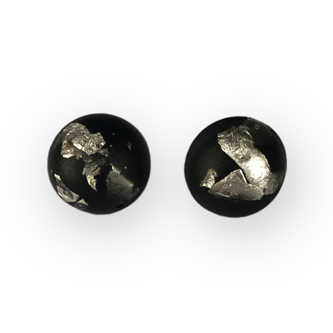 Two black spherical beads with irregular silver foil-like patterns on their surfaces are positioned against a white background. The beads, featured as part of our Luxe Stud Earrings Black by Angela O'Keefe collection, have a smooth and glossy finish, showcasing an artistic and contrasting design.