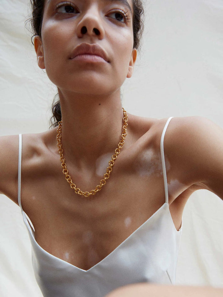 A person with a calm expression on their face is wearing a white spaghetti-strap top and a delicate, 22 carat Mayal Gold Chain Necklace by Shyla. The person has vitiligo, visible as light patches on their skin, and is positioned against a plain, light-colored background.