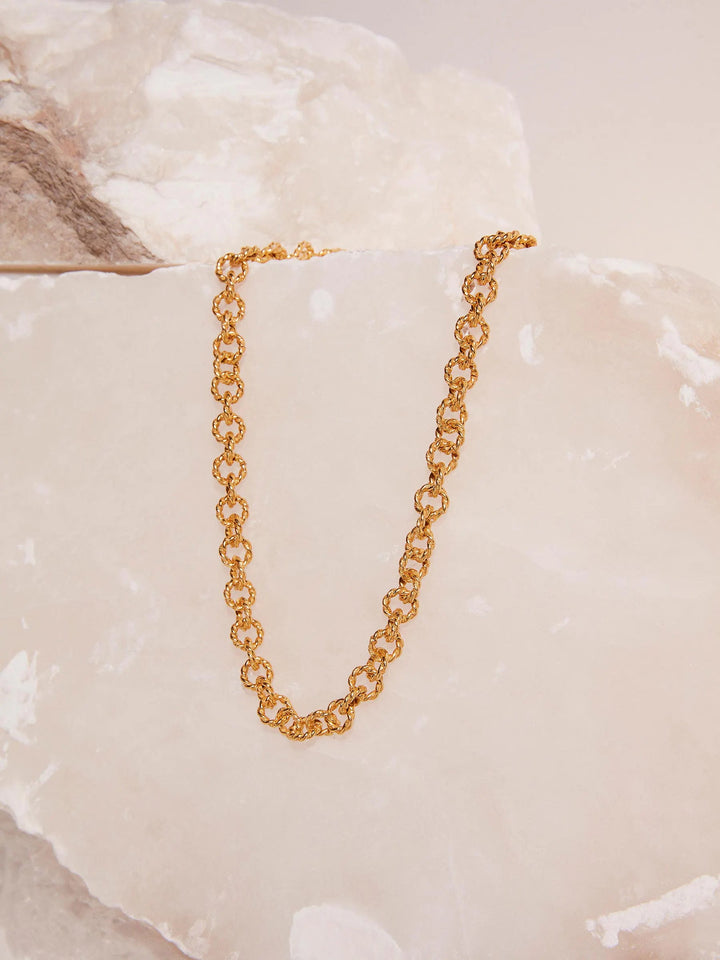 A Mayal Gold Chain Necklace by Shyla is elegantly displayed against a backdrop of light beige and cream-colored marble slabs. The 22-carat piece has a shiny finish, and its intricate design stands out beautifully against the smooth, natural stone surface.