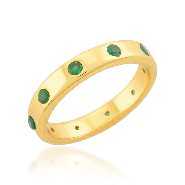 The Shyla Mela Ring, crafted from 22 carat gold, is adorned with small, round green gemstones evenly spaced around the polished band. The background is white, highlighting the ring's details and shine while adding a touch of color and elegance to the design.