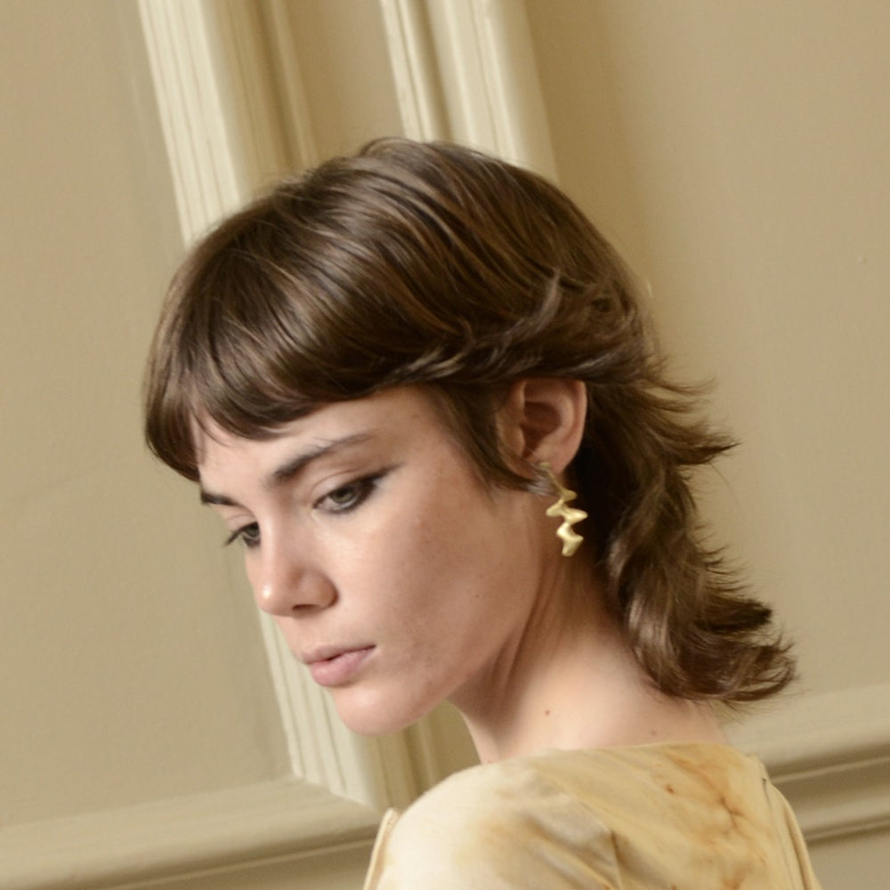 A person with short, layered brown hair, winged eyeliner, and a light top is shown in profile wearing the Blaze Studs by Madeleine Sculptural Jewellery—sculptural earrings crafted from recycled brass—against a beige wall with decorative molding.