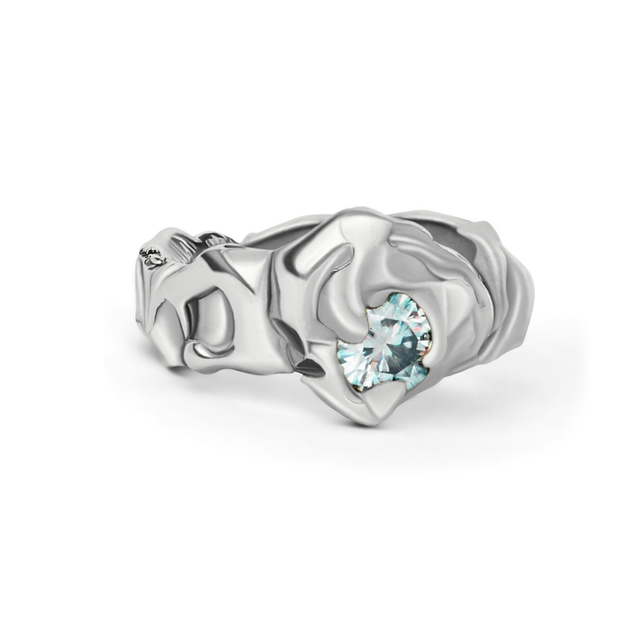 The Molten Ring with Moissanite by Madeleine Sculptural Jewellery showcases a silver band with an abstract rose design and a round, sparkling light blue gemstone for sustainable style and timeless elegance.