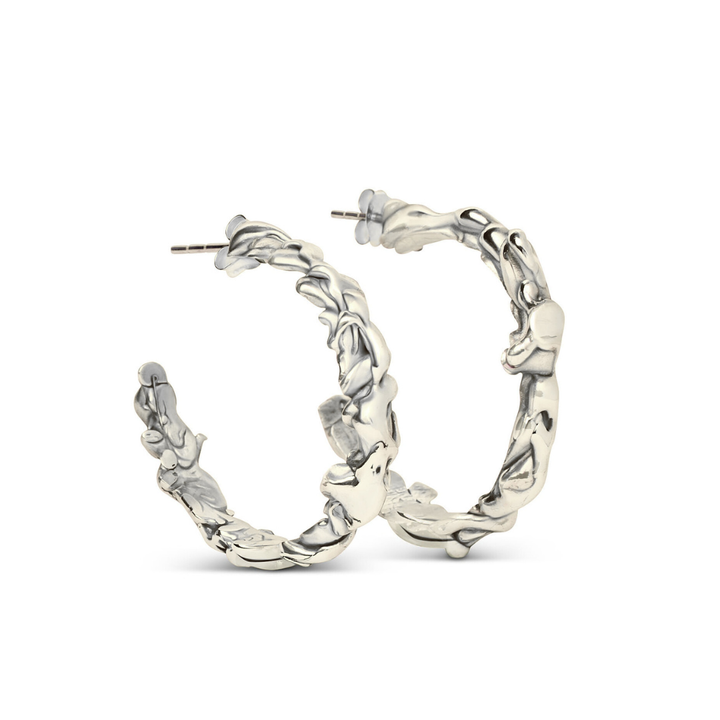 The Molten Hoops Large by Madeleine Sculptural Jewellery are silver hoop earrings with a textured, wavy design, reminiscent of hand-carved pieces, displayed upright against a white background.