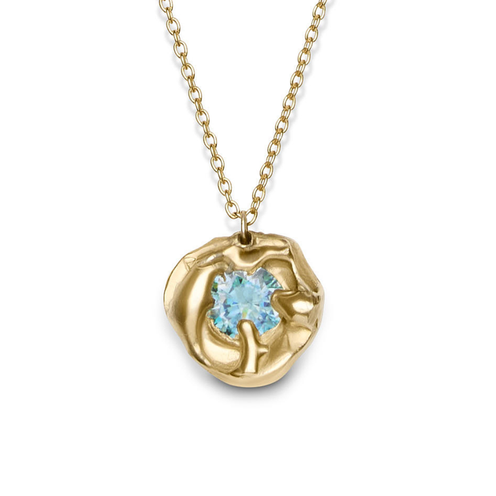 The Molten Necklace with Moissanite by Madeleine Sculptural Jewellery features a hand-carved, organic-shaped gold pendant in recycled brass, showcasing a round light blue moissanite—a stunning, sustainable statement piece.
