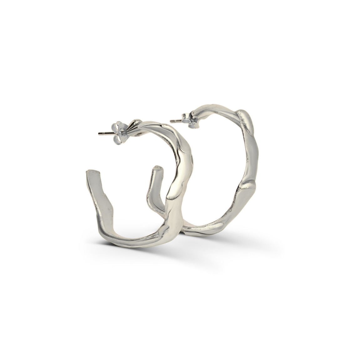 The Liana Hoops by Madeleine Sculptural Jewellery are silver earrings featuring an organic, wavy, hand-carved design, displayed standing upright against a white background.
