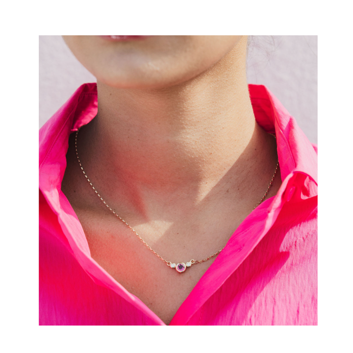 Close-up of a person wearing a bright pink shirt and the Madeleine Fine Jewellery Rose Embrace 9ct Yellow Gold Necklace with Two Diamonds—a delicate gold necklace featuring a small round pendant set with two diamonds—against a light background.