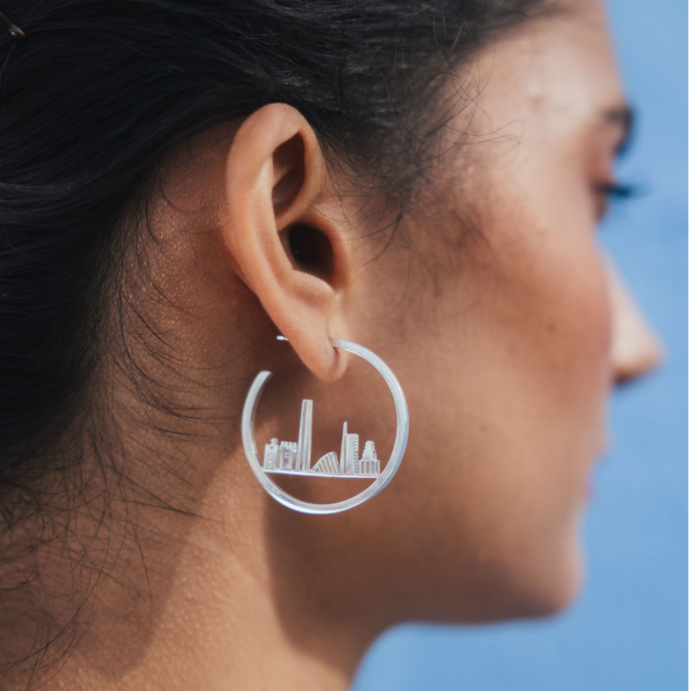 Close-up of a person’s ear wearing a large Horizon of Dublin Hoops earring by The Collective Dublin, featuring a city skyline design. This handcrafted jewellery piece highlights sustainable craftsmanship, set against a blurred blue background.
