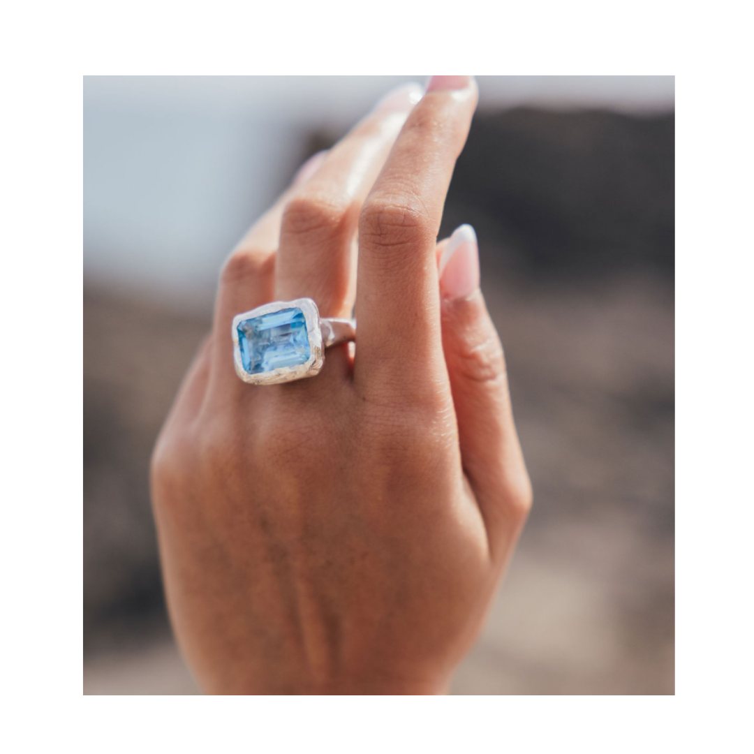 A hand with manicured nails showcases the Glazier Ring by Madeleine Sculptural Jewellery—a large, rectangular blue gemstone set in silver—held up against a blurred outdoor background.