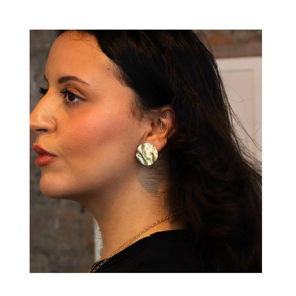 A woman with long dark hair is shown in profile, wearing Madeleine Sculptural Jewellery’s Tide Studs Large—round, iridescent recycled brass earrings. She has a neutral expression and wears a black top against a blurred background.