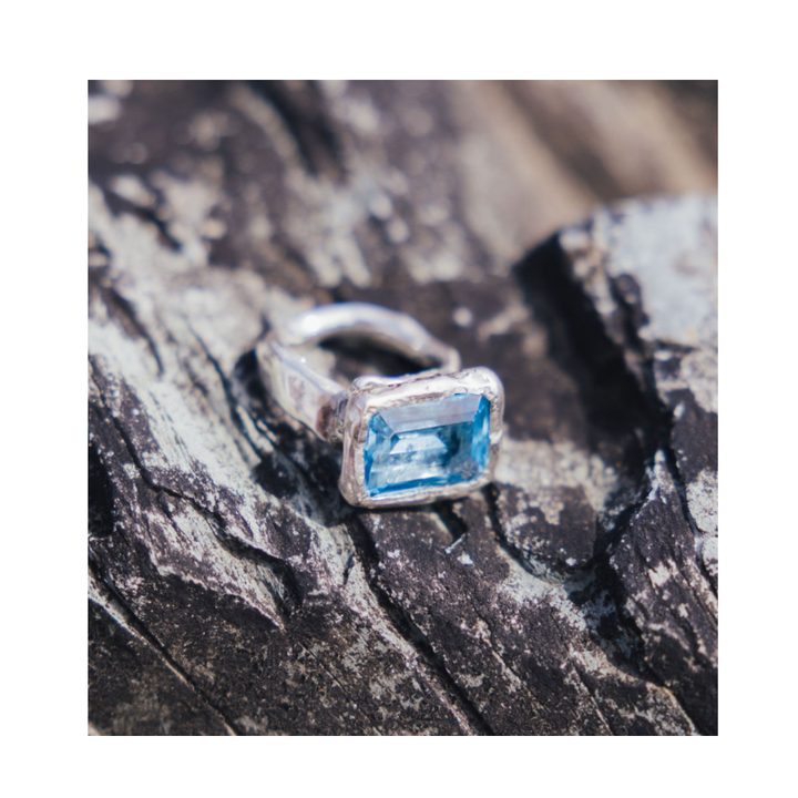 The Glazier Ring by Madeleine Sculptural Jewellery, featuring a rectangular blue gemstone set in silver, is displayed on a rough, textured stone surface.