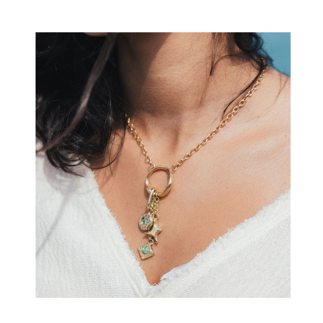 A woman wearing the Spark Necklace by Madeleine Sculptural Jewellery—a gold chain necklace featuring a round clasp, star charm, and gemstone pendants—paired with a white textured top with a wide neckline.