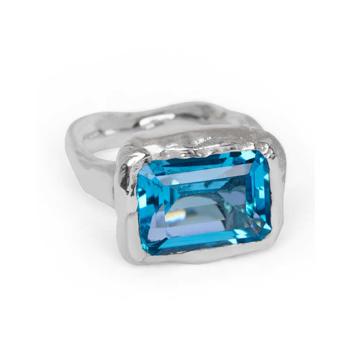 The Glazier Ring by Madeleine Sculptural Jewellery features a large, rectangular, faceted blue gemstone set in a thick, organic-style silver bezel and is displayed on a white background.