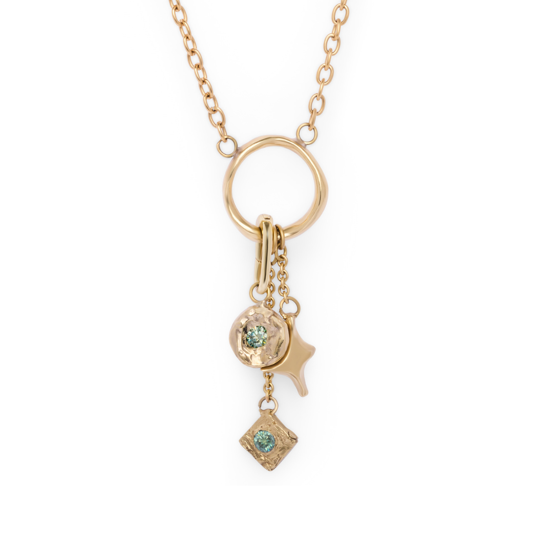 The Spark Necklace from Madeleine Sculptural Jewellery features a gold chain with a circular pendant, adorned with three dangling charms: a textured disc set with a green gem, an abstract star shape, and a small square charm accented by a green stone. Displayed against a white background.