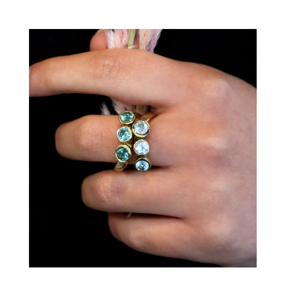 A hand wearing two Madeleine Sculptural Jewellery Moonwater Rings, each with three round blue gemstones, gently holds a light pink feather against a black background, highlighting the elegance of sustainable design.