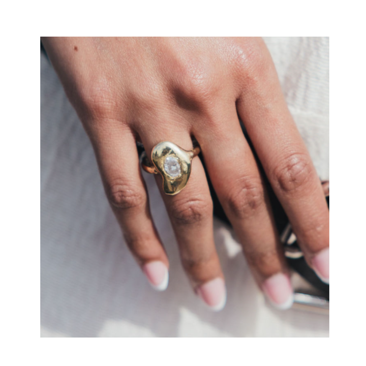 A hand adorned with the Carraig Ring from Madeleine Sculptural Jewellery—with its gold band and clear stone centerpiece—rests gracefully on a white fabric surface. The nails are manicured with a natural pink base and white tips, creating an elegant look ideal for a one-off special occasion.