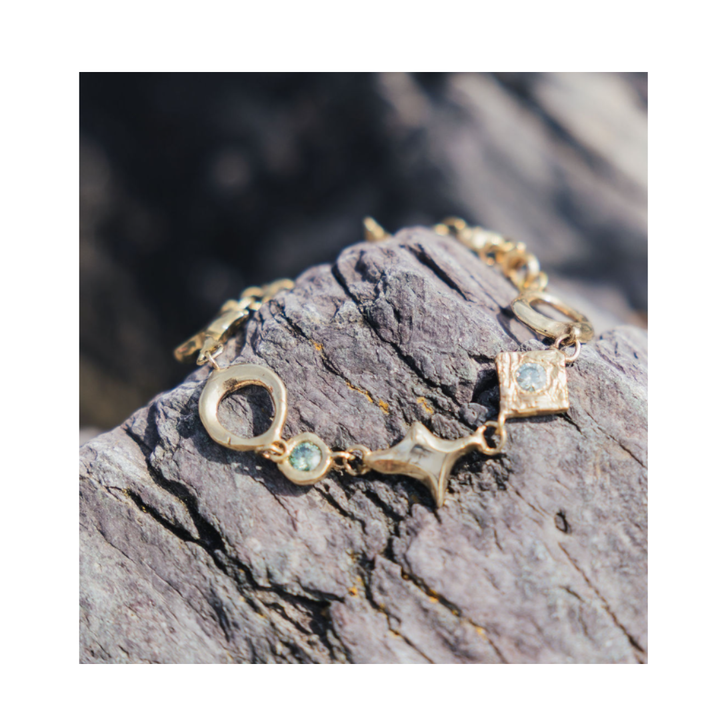 The Spark Bracelet in Brass by Madeleine Sculptural Jewellery, featuring distinctive geometric charms and small blue gemstones, rests on a textured gray rock in natural light.