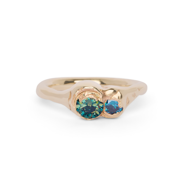 The Dune Ring by Madeleine Sculptural Jewellery features an organic, wavy gold band with two round, blue-green gemstones set side by side in an asymmetrical bezel-style setting.