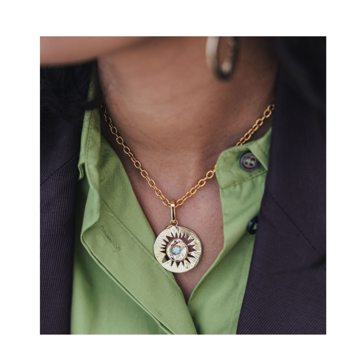 A person wearing the Tundra Necklace by Madeleine Sculptural Jewellery—a gold chain necklace with a large, round sunburst pendant—paired with a green button-up shirt under a dark blazer.