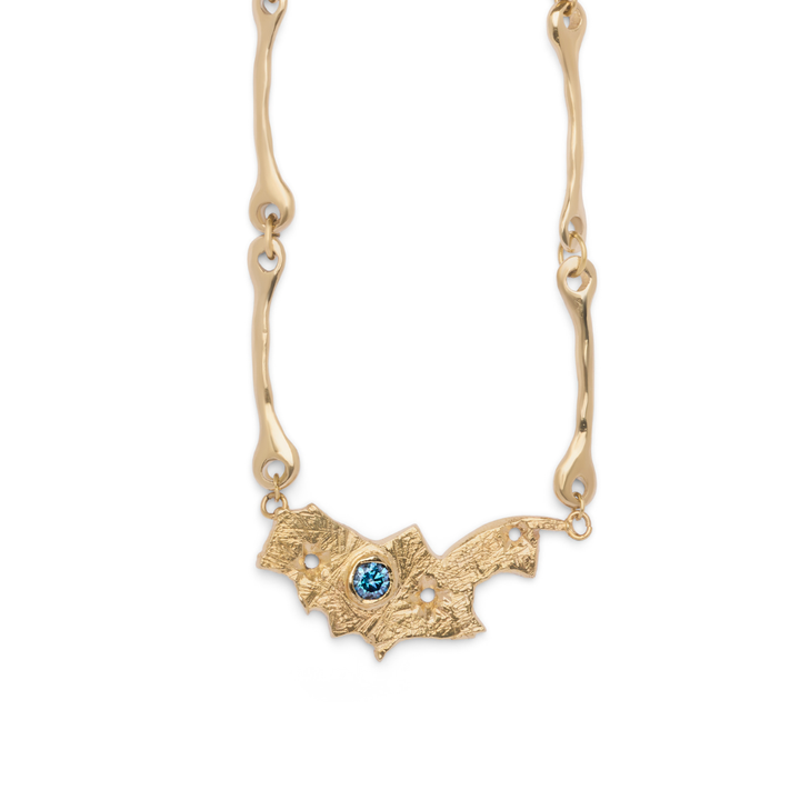 The Blaze Necklace by Madeleine Sculptural Jewellery features a bat-shaped pendant with a textured surface, adorned with small pearl accents and a striking central blue gemstone. Its chain is designed with organic, wavy links. Photographed on a white background.