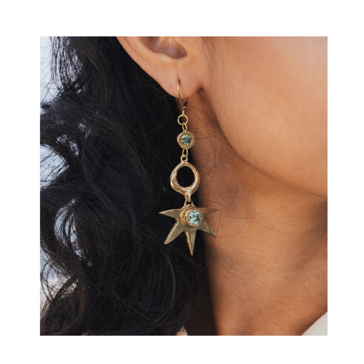 A close-up of a person with long, wavy dark hair wearing the Réalta Earrings with 2 moissanite by Madeleine Sculptural Jewellery—a striking 3-part gold earring adorned with a moon and star design, featuring a round gem at the top and a sparkling star-shaped charm at the bottom.