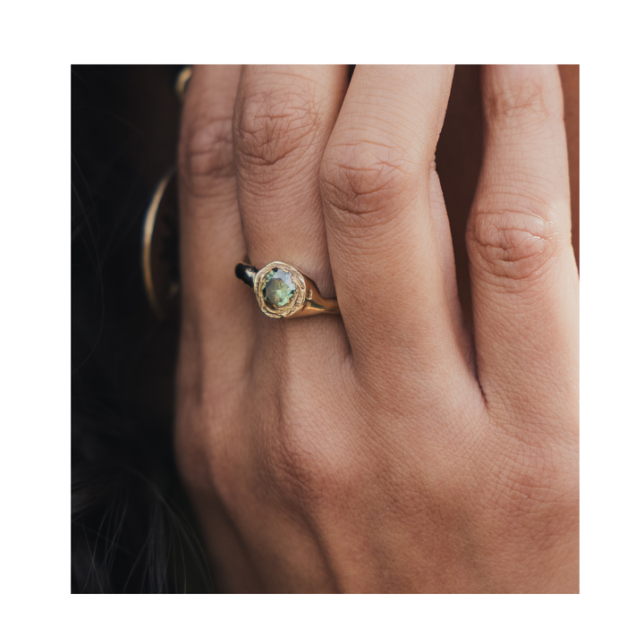 A close-up showcases a hand wearing the Dawn Ring by Madeleine Sculptural Jewellery—a gold band featuring a green gemstone—evoking an aura of serenity and simplicity. Worn on the middle finger, the hand rests gracefully on a dark surface, and the image is tastefully framed with a white border.