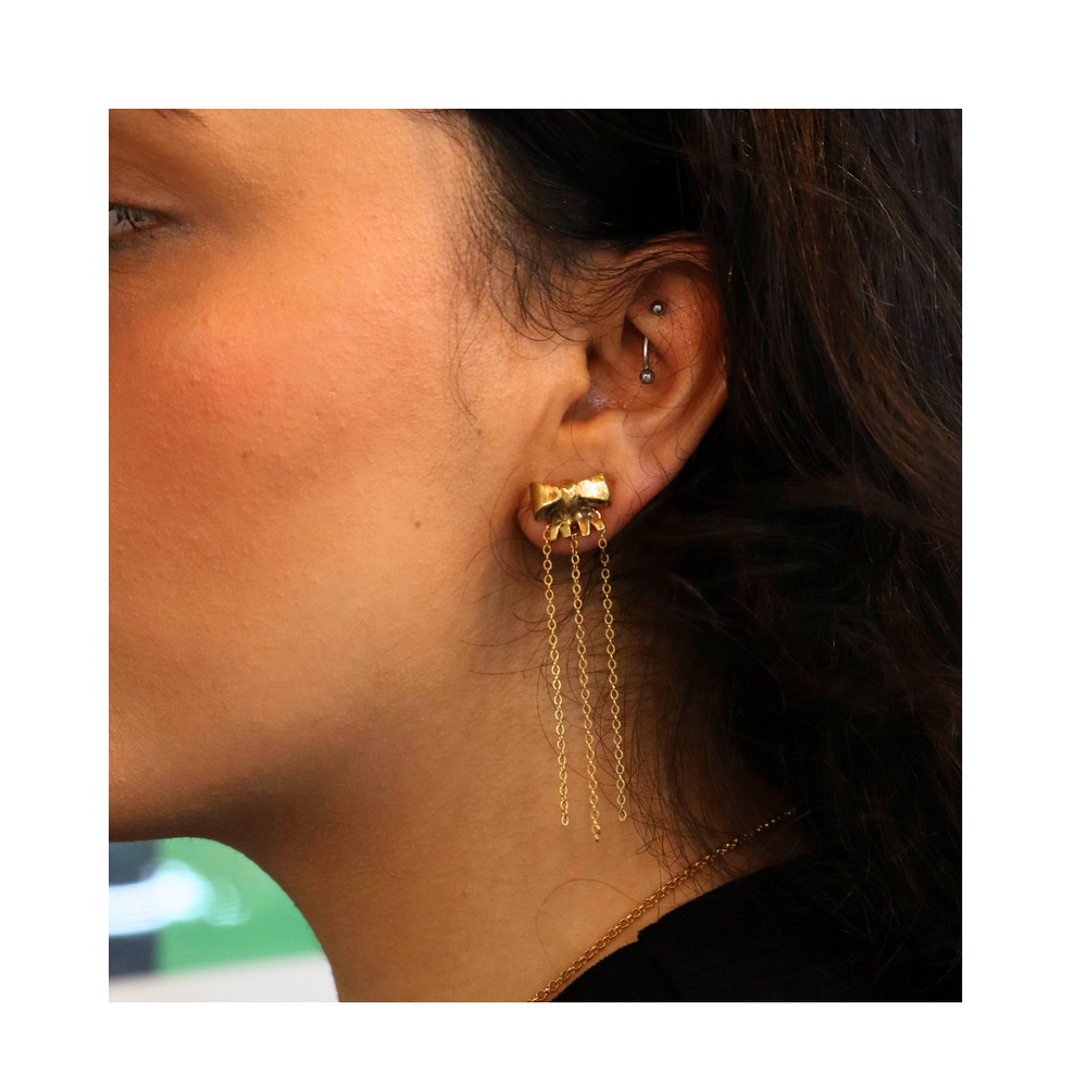 Close-up of a woman's ear wearing Bow earrings by Madeleine Sculptural Jewellery, featuring gold plated brass with a rectangular stud and three delicate chain dangles—an elegant, sustainable jewellery piece for multiple piercings.