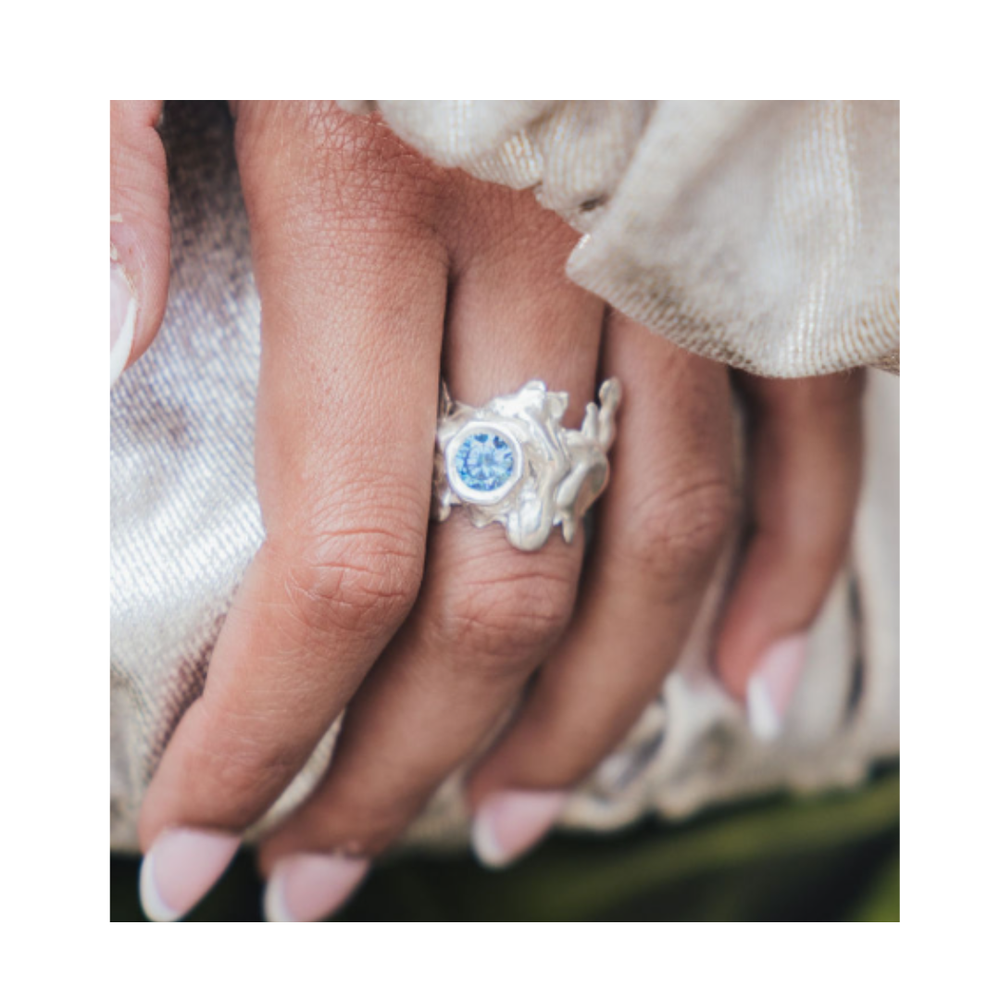 A close-up of a hand with manicured nails showcases the Solstice Ring with Large Moissanite by Madeleine Sculptural Jewellery. This striking example of custom design, set with a brilliant pale blue moissanite, rests elegantly on light, shimmery fabric—reflecting the allure of unique pieces made to order.