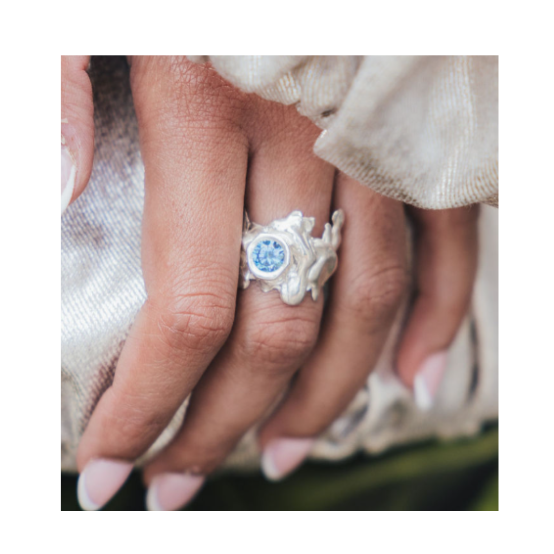 A close-up of a hand with manicured nails showcases the Solstice Ring with Large Moissanite by Madeleine Sculptural Jewellery. This striking example of custom design, set with a brilliant pale blue moissanite, rests elegantly on light, shimmery fabric—reflecting the allure of unique pieces made to order.