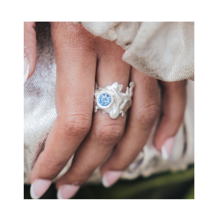 A close-up of a hand with manicured nails showcases the Solstice Ring with Large Moissanite by Madeleine Sculptural Jewellery. This striking example of custom design, set with a brilliant pale blue moissanite, rests elegantly on light, shimmery fabric—reflecting the allure of unique pieces made to order.