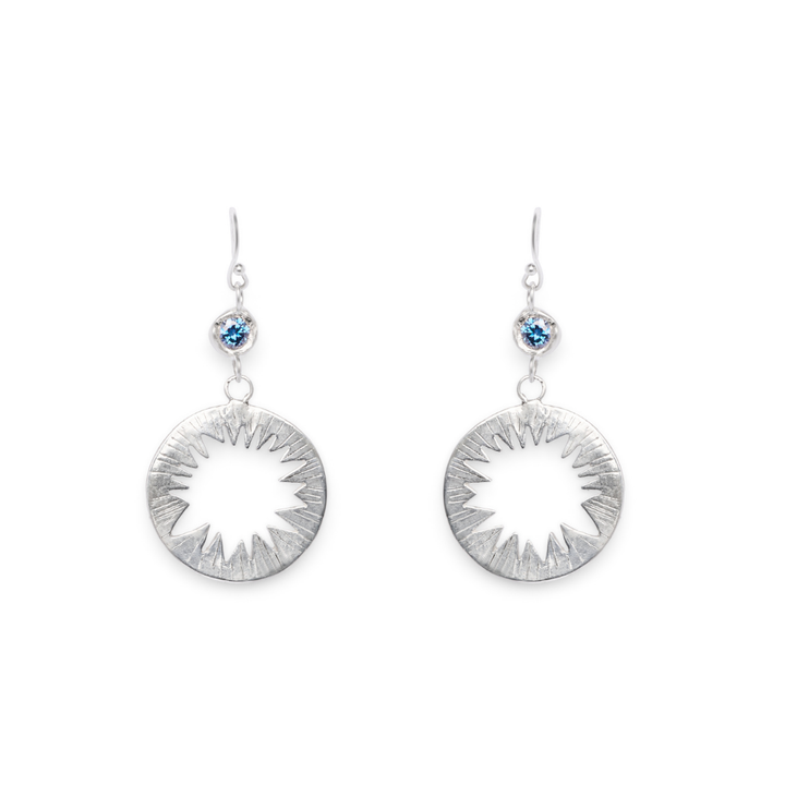 The Tundra Earrings with Moissanite by Madeleine Sculptural Jewellery feature blue moissanite gemstones set at the top and circular sunburst cutout designs dangling below on hooks, displayed against a white background.