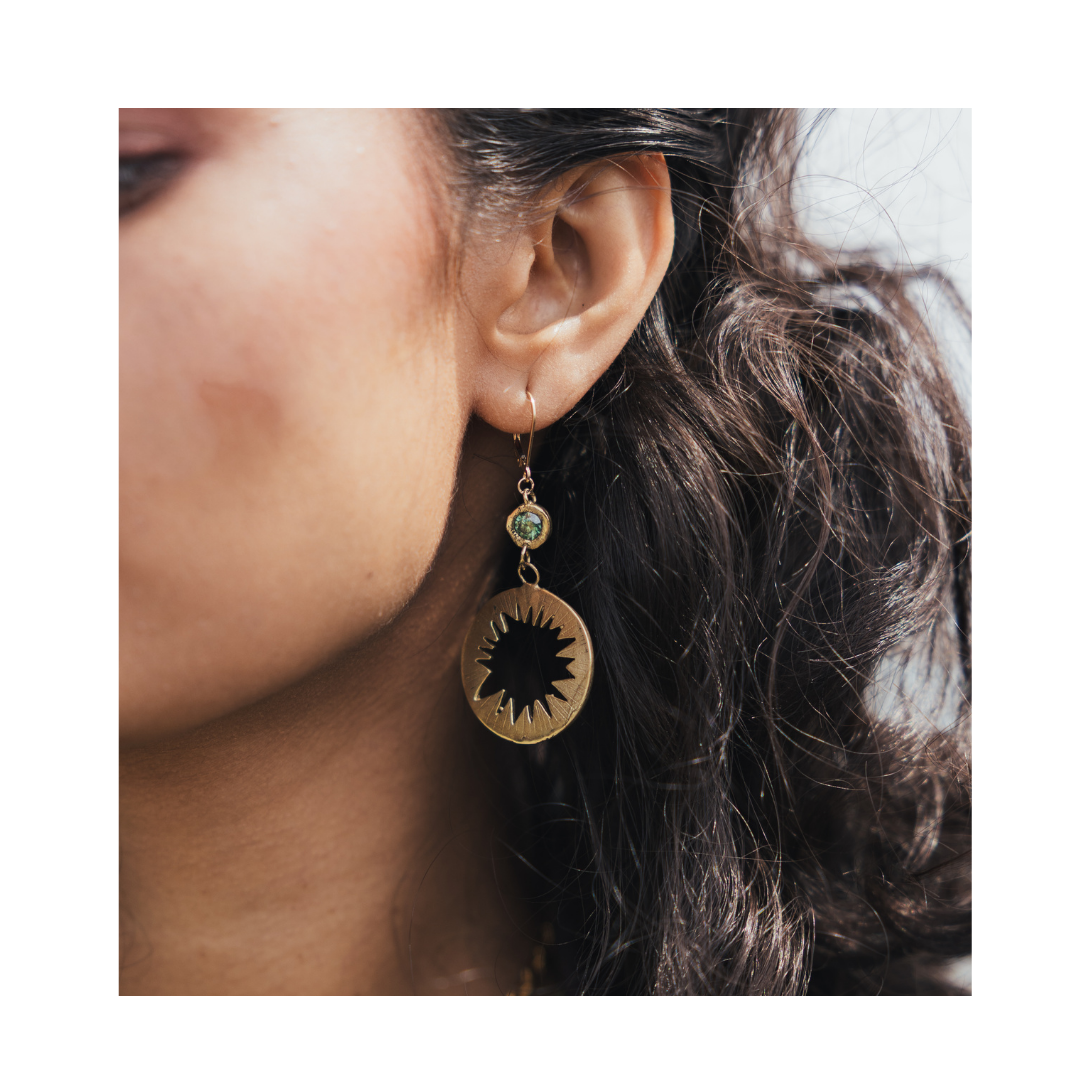 Close-up of a woman’s ear with wavy dark hair, wearing the Madeleine Sculptural Jewellery Tundra Earrings with Moissanite—large, round gold earrings featuring a black starburst design and a small green bead above it.