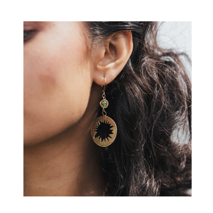 Close-up of a woman’s ear with wavy dark hair, wearing the Madeleine Sculptural Jewellery Tundra Earrings with Moissanite—large, round gold earrings featuring a black starburst design and a small green bead above it.
