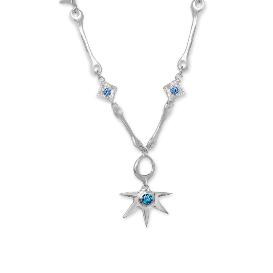A silver “Bone Necklace with Star Centre” by Madeleine Sculptural Jewellery, adorned with blue gemstones and featuring star-shaped and diamond-shaped pendants, including a central blue gem set in an eight-pointed star.