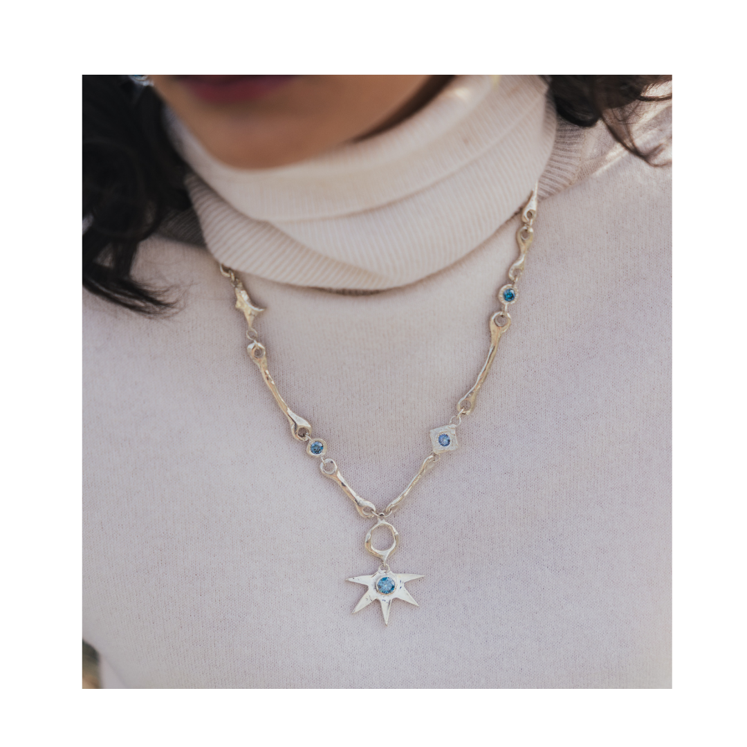A person wearing a cream turtleneck sweater and the Madeleine Sculptural Jewellery Bone Necklace with Star Centre, featuring silver links, blue gemstones, and a large star-shaped pendant.